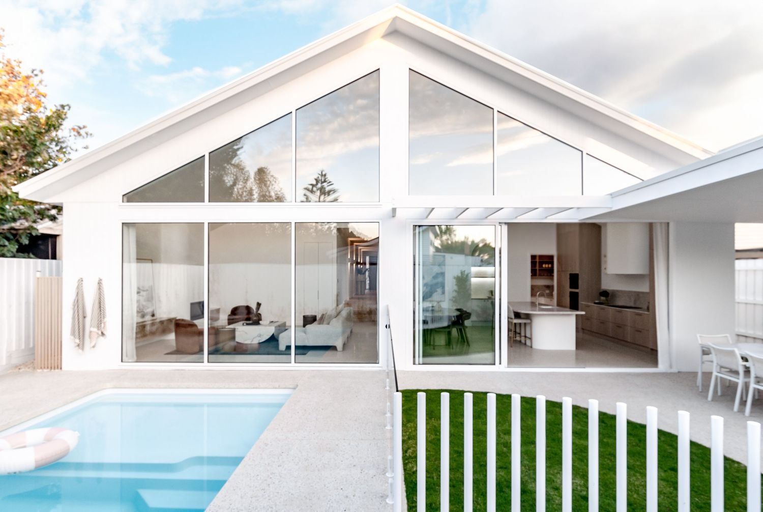 Modern white house with large glass windows, gable roof, swimming pool and manicured lawn in the backyard.