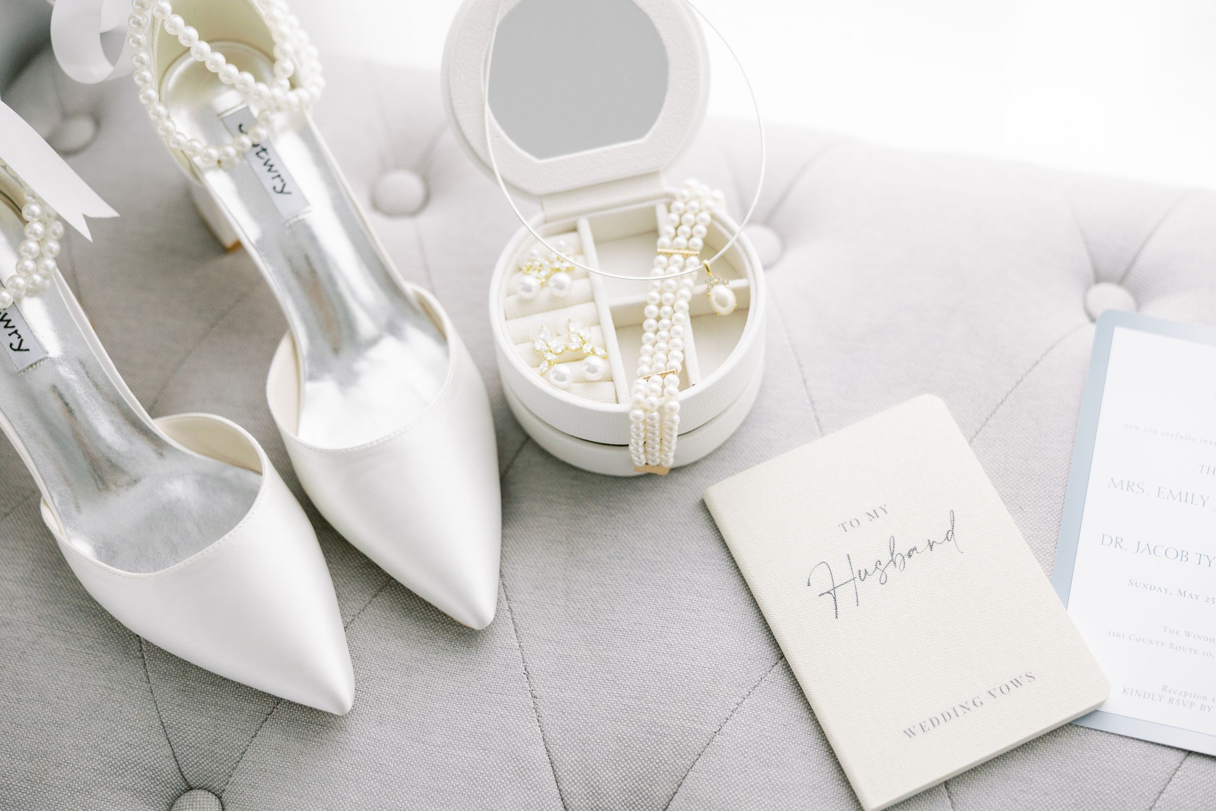 White bridal high heels next to pearl hair accessories and a wedding invitation on a light gray surface.