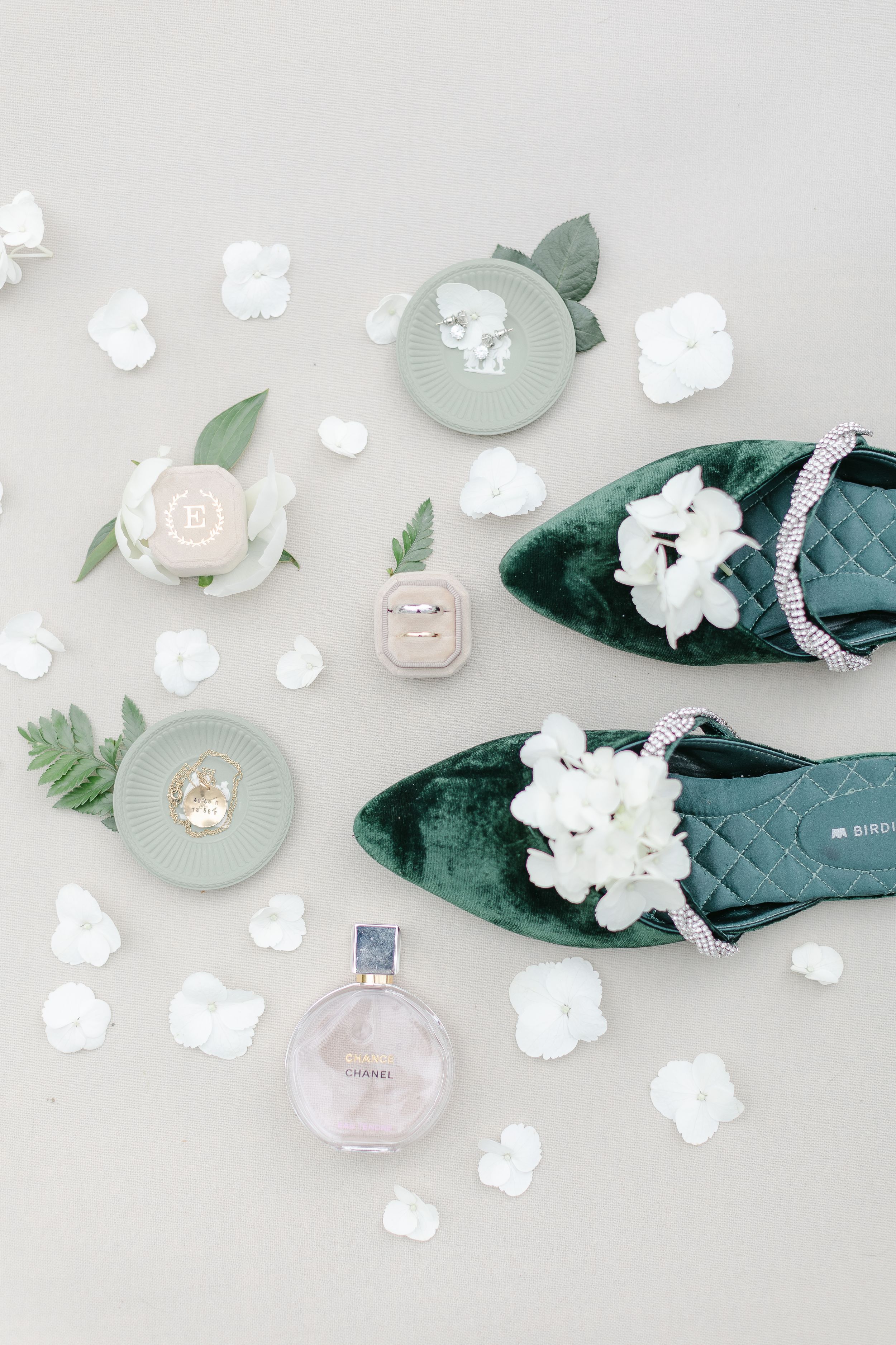 Elegant green velvet flats with white flowers displayed alongside perfume bottles and delicate petals on a light gray surface.