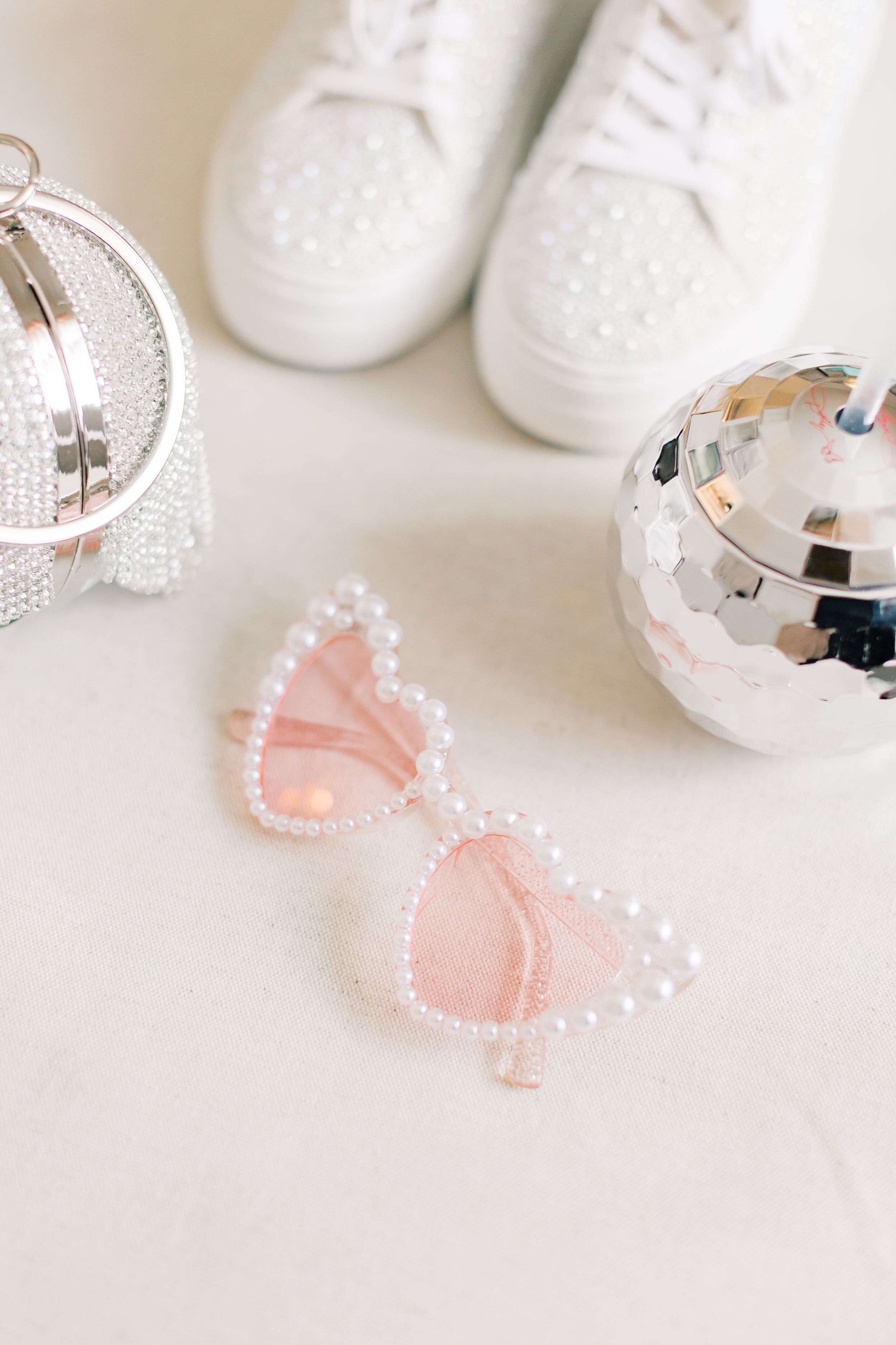 Pink pearl-rimmed sunglasses with white sneakers and disco balls on light background create a festive party styling setup.