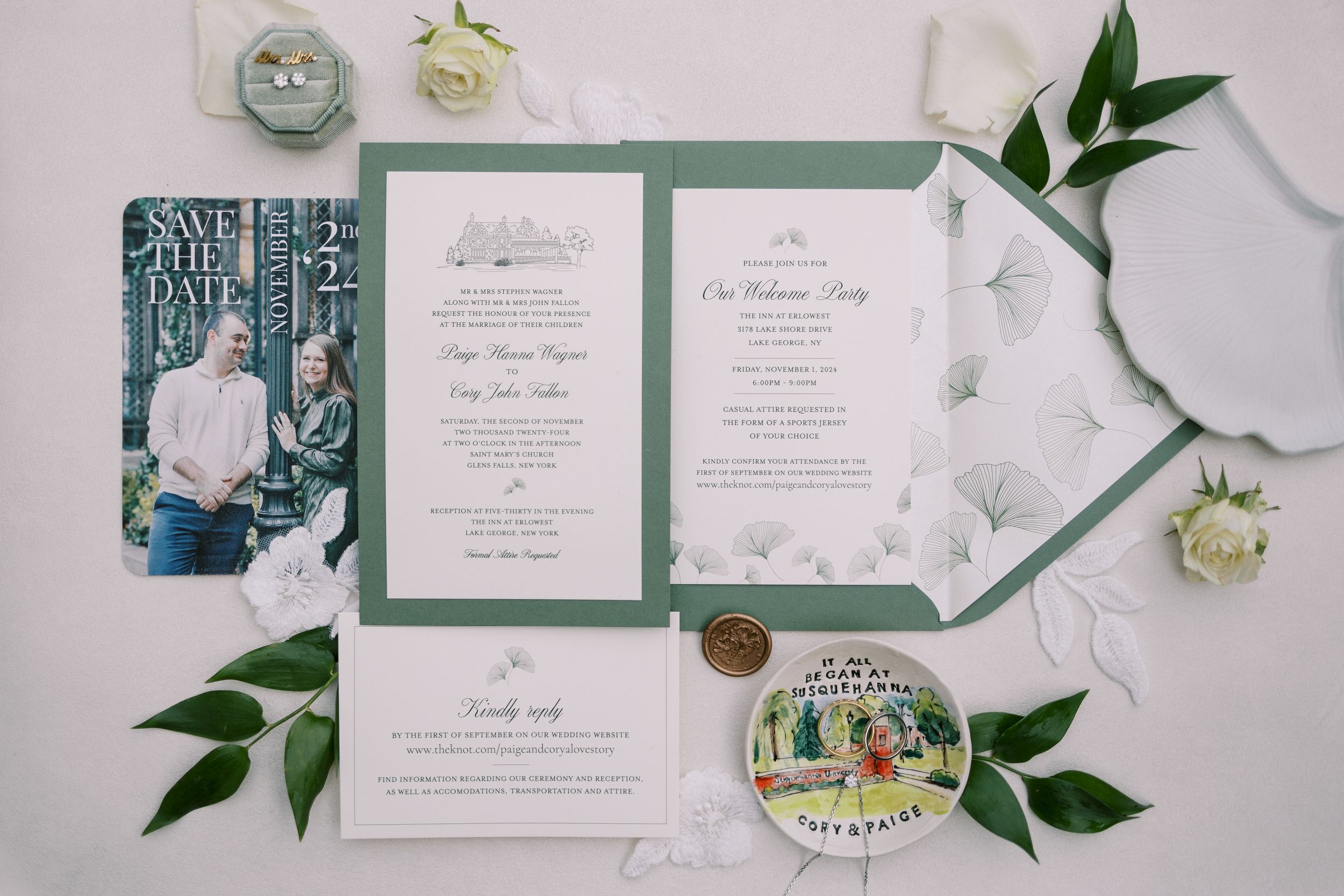 Elegant white and sage green wedding invitation suite with delicate floral details and greenery accents laid on white surface.