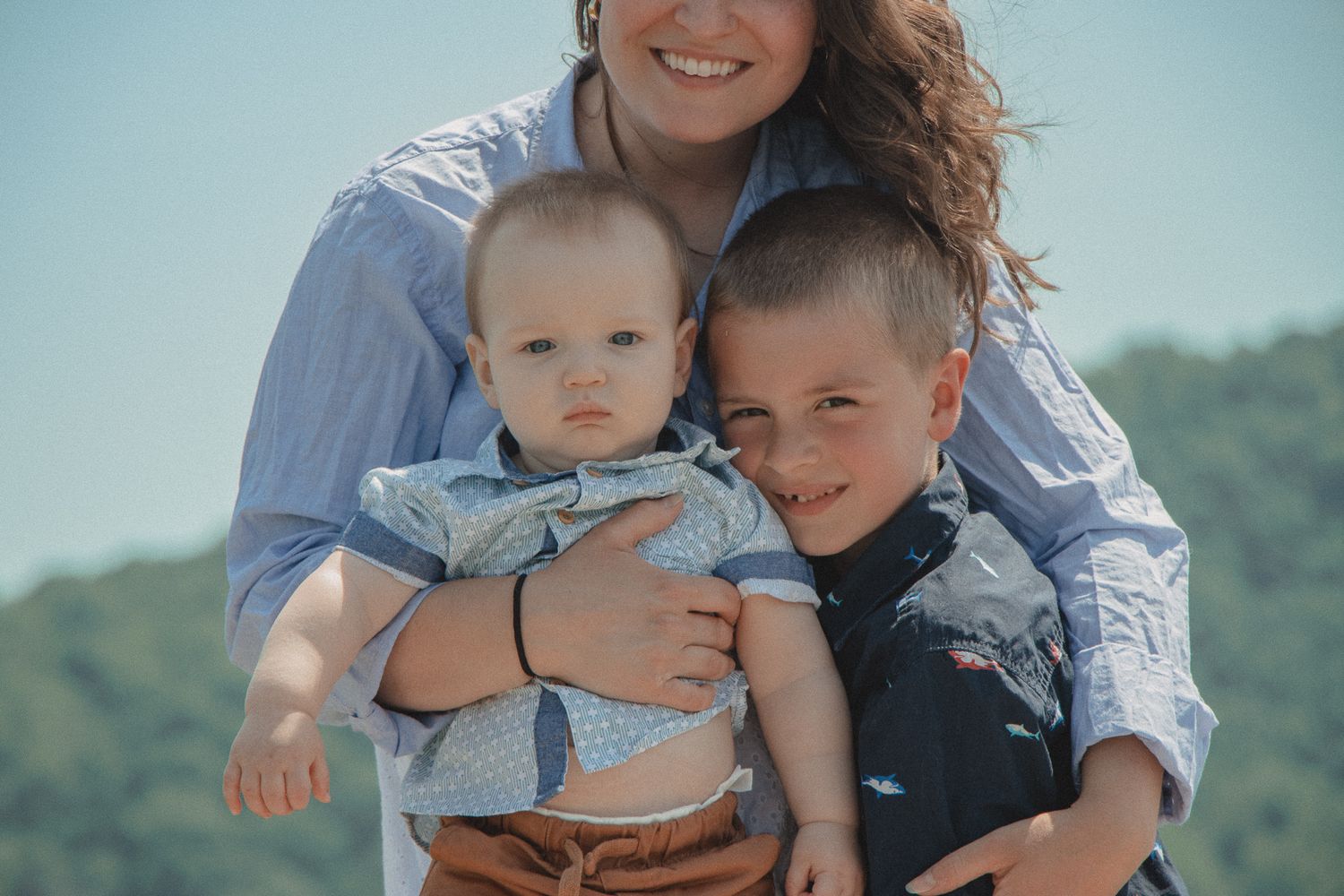 Mom hugs her baby and young son, all smiling together outdoors with soft mountain views in the background