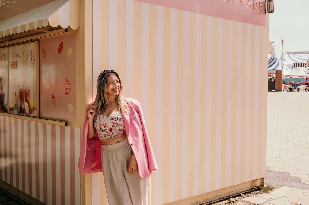 Person in pink blazer and floral top stands against striped wall on sunny day.