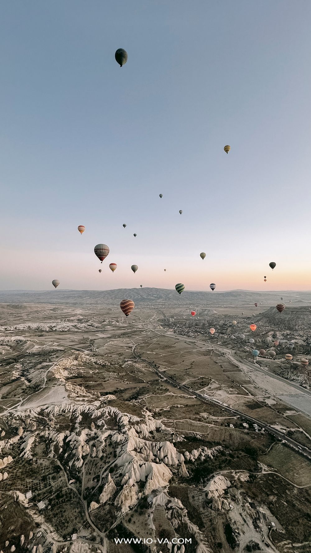 Aerial view of numerous hot air balloons floating over Cappadocia's rocky terrain at sunrise.