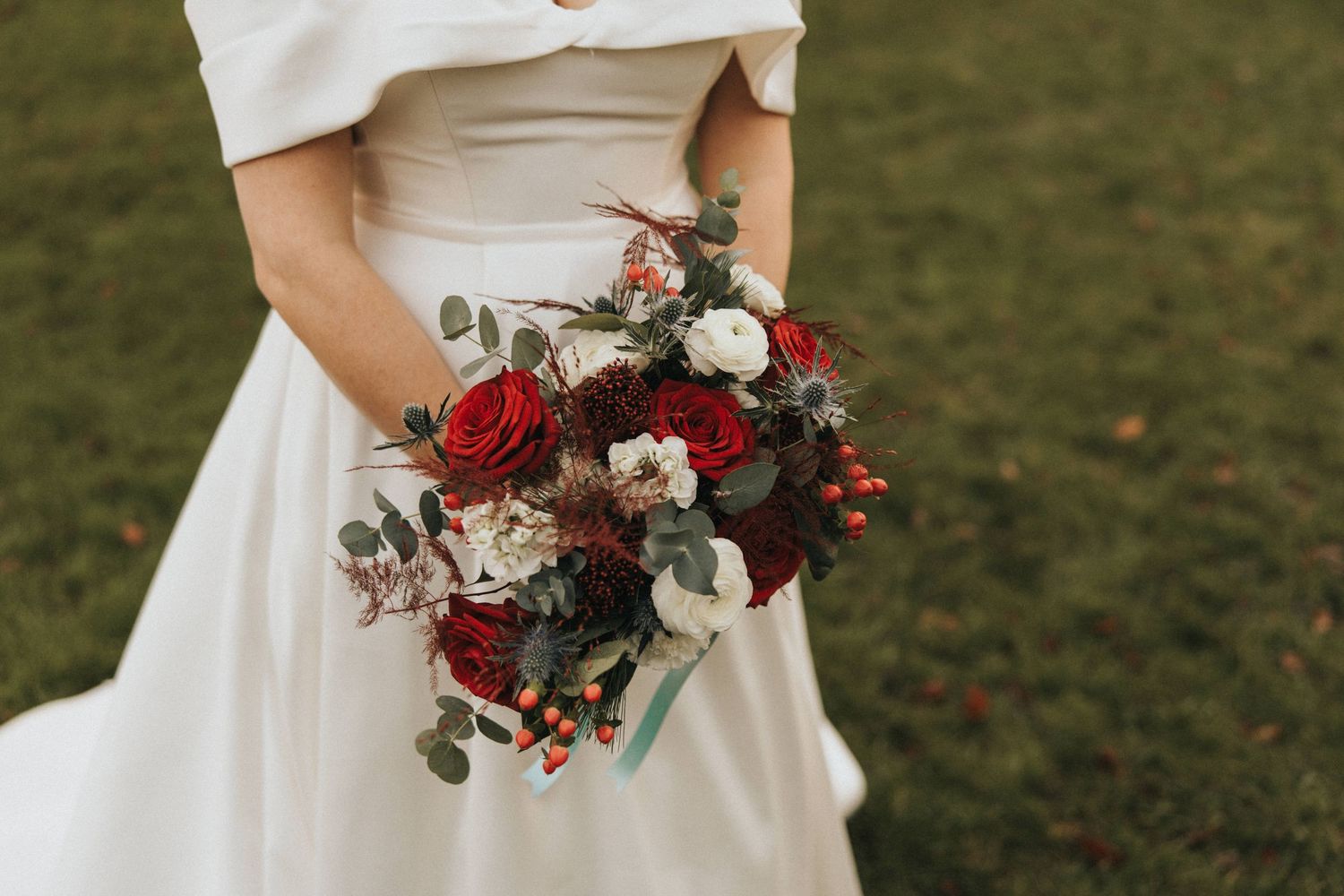 Bride in elegant white dress holds a romantic burgundy and cream floral bouquet with eucalyptus against a grassy background.