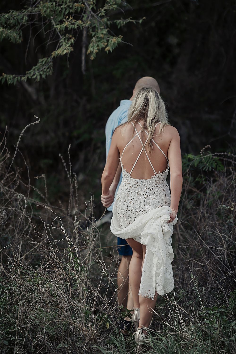 A couple walking through a wooded area, with the woman wearing a white lace dress with crossed back straps.