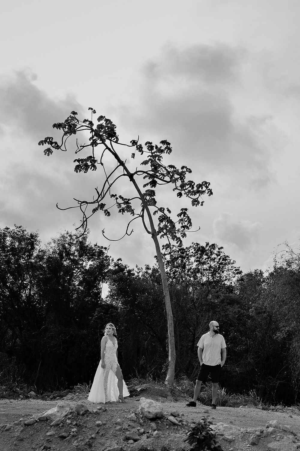 A monochromatic artistic photo of a couple standing apart beneath a twisted tree on a hilltop against a cloudy sky.