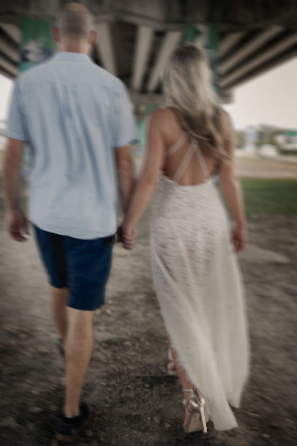 A couple walks hand in hand under a bridge, with the person in white wearing a flowing maxi dress.