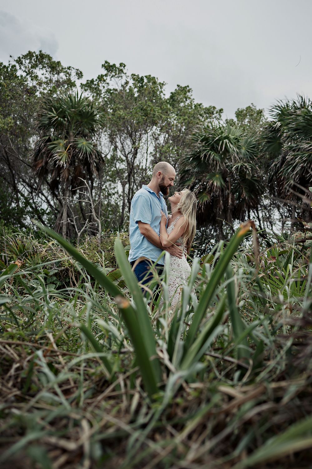 A couple shares an intimate moment surrounded by lush tropical plants and palm trees in a natural outdoor setting.