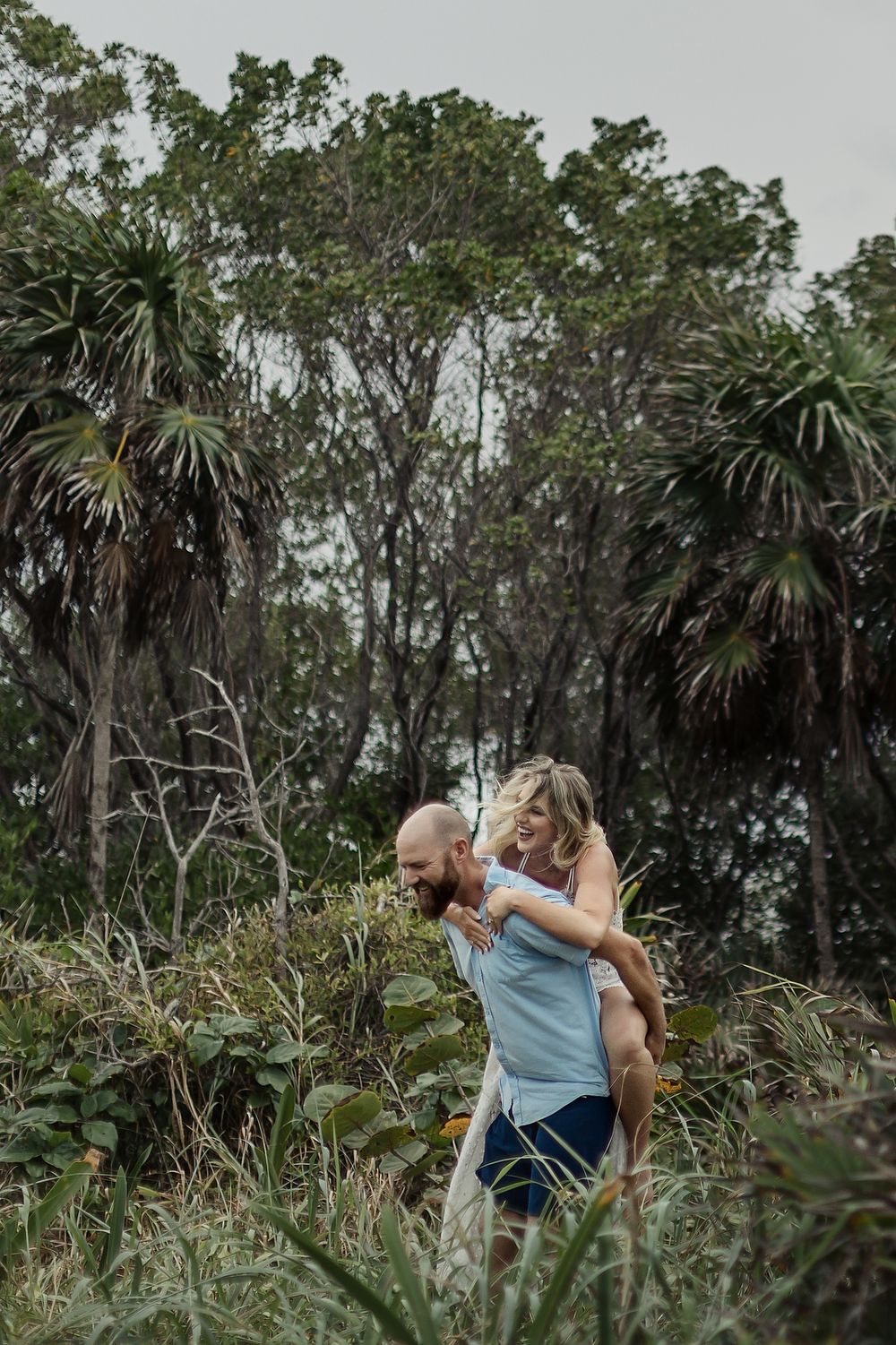 A couple embraces among palm trees and lush tropical foliage in a moody outdoor setting.