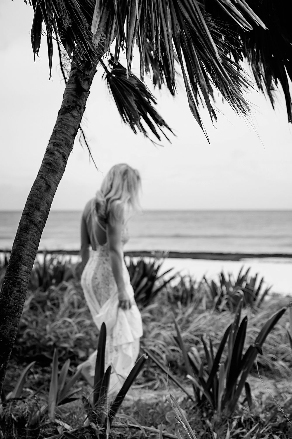 Black and white scenic view of tropical palm trees and oceanfront beach with figure in flowing white dress at sunset.
