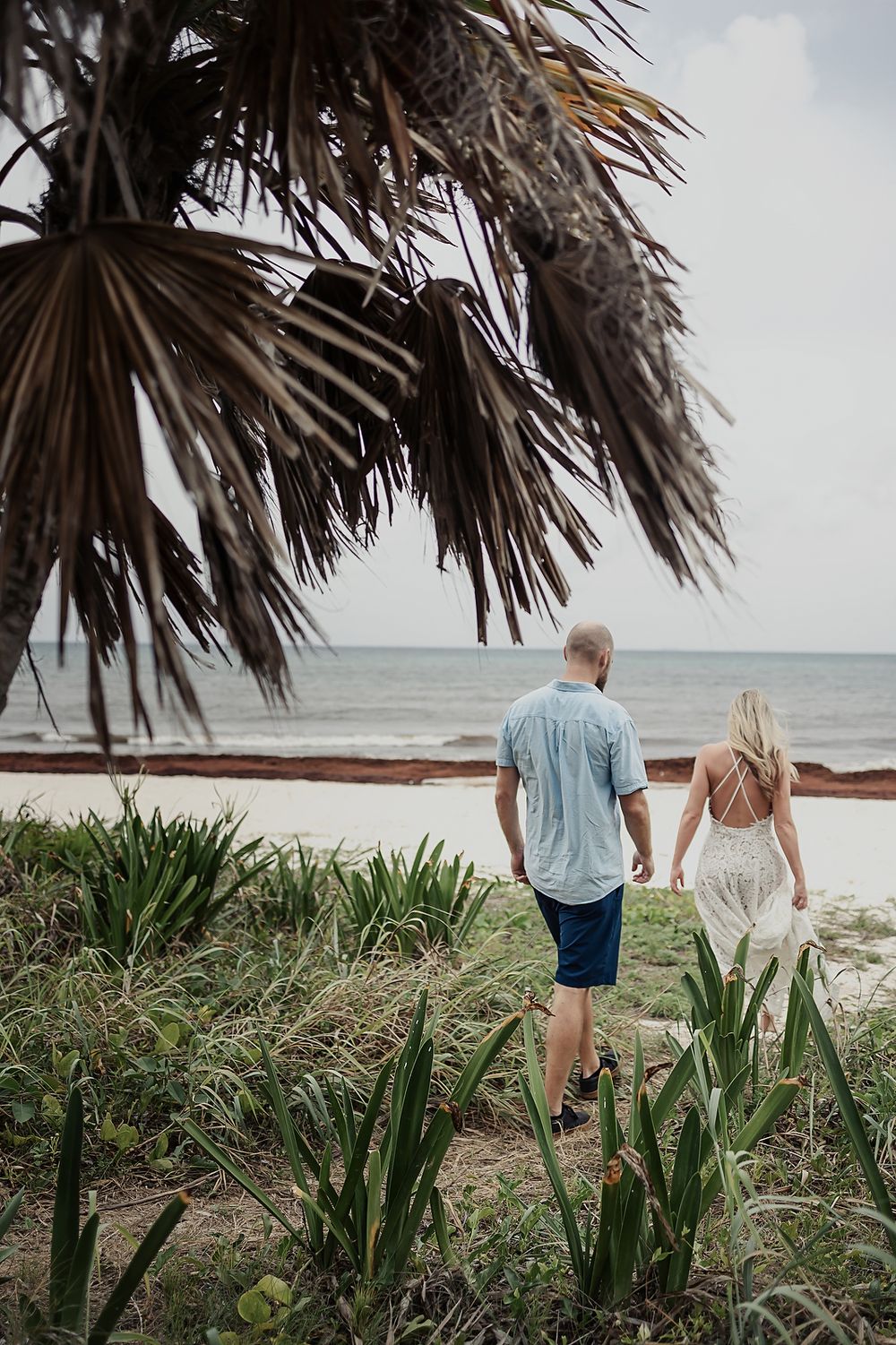 A couple stands together overlooking a tropical beach with palm fronds and sea grass in the foreground.