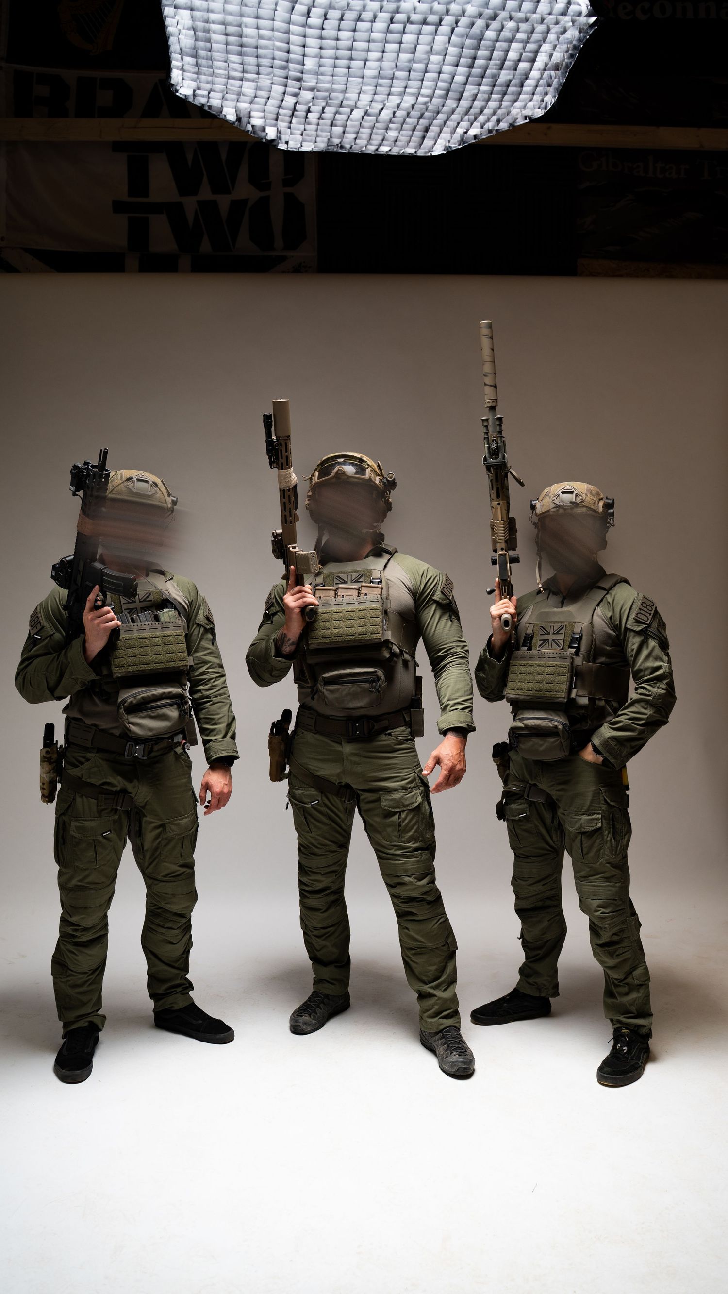 Three uniformed soldiers in tactical gear and combat vests pose with rifles in a studio setting against a white background.