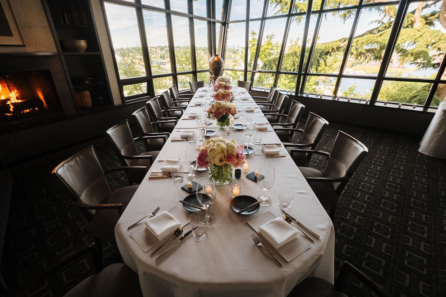 A long white dining table set for a formal event by floor-to-ceiling windows with a fireplace and mountain views.