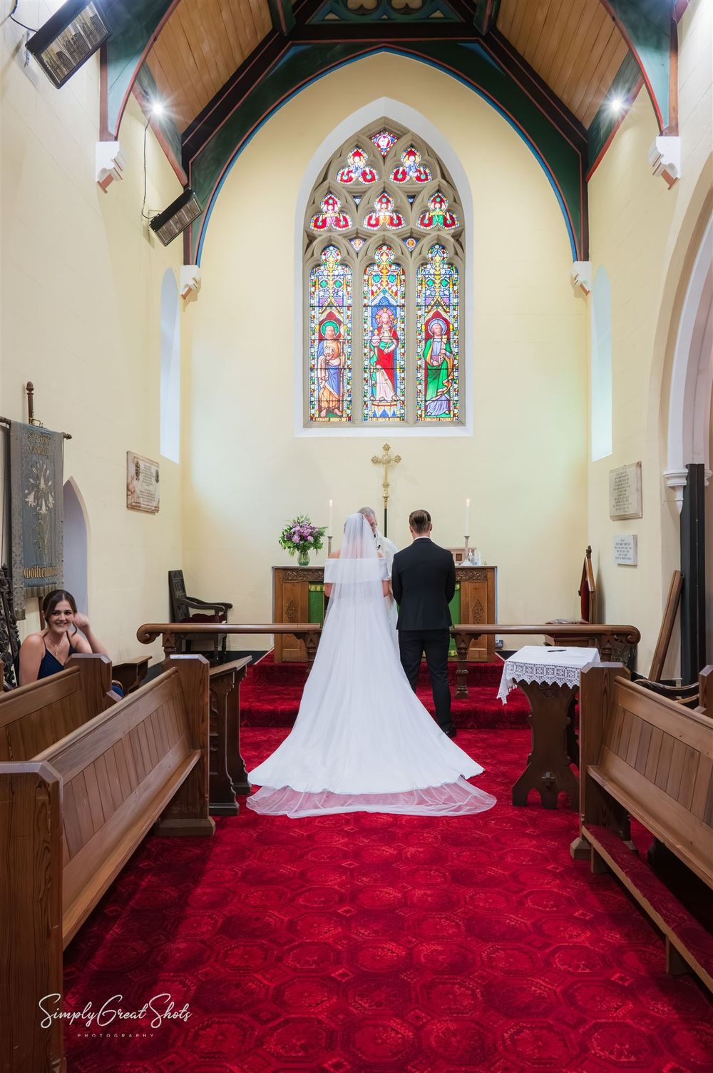 A bride and groom stand before a beautiful stained glass window in St Peters Church, Kidderminster, with a red carpet and wooden pews.