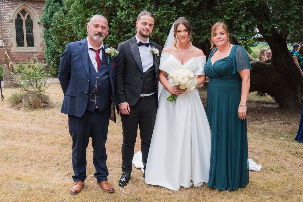 A formal wedding portrait group standing outside a St Peters church in Kidderminster with gothic windows in a natural garden setting.