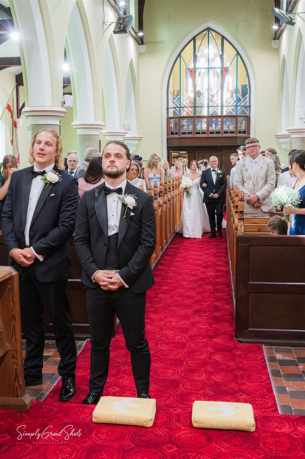 A groom waits patiently as his bride walks down the aisle in a traditional church with gothic arched windows and wooden pews.