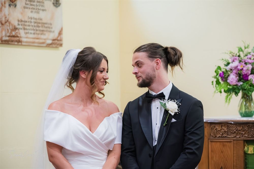 A couple exchange loving glances during their wedding ceremony with floral arrangements visible in the background.