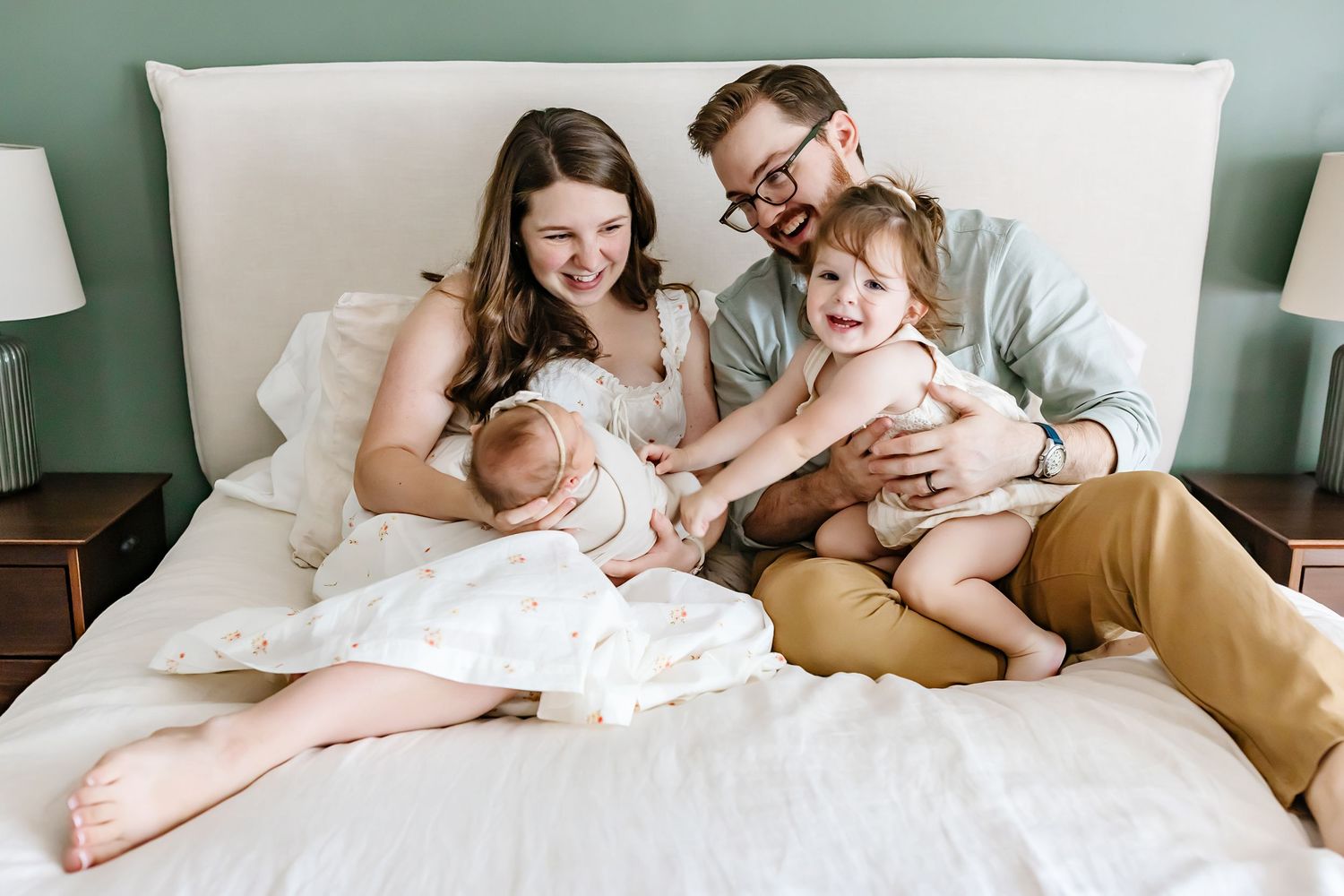 A family relaxes together on a white bed against a green wall in a candid lifestyle portrait.