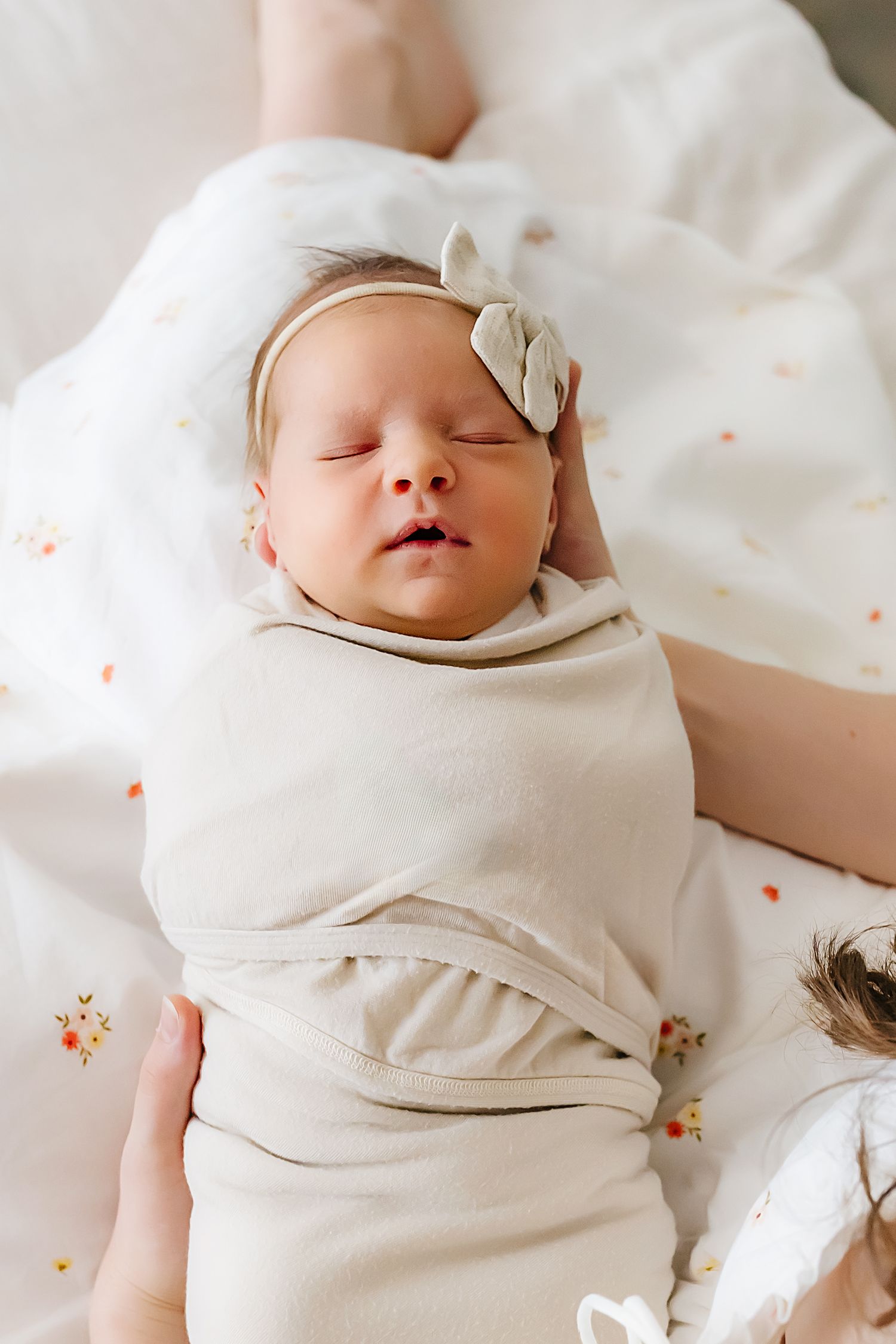 A sleeping newborn baby in cream colored wrap and bow headband rests peacefully on floral bedding.