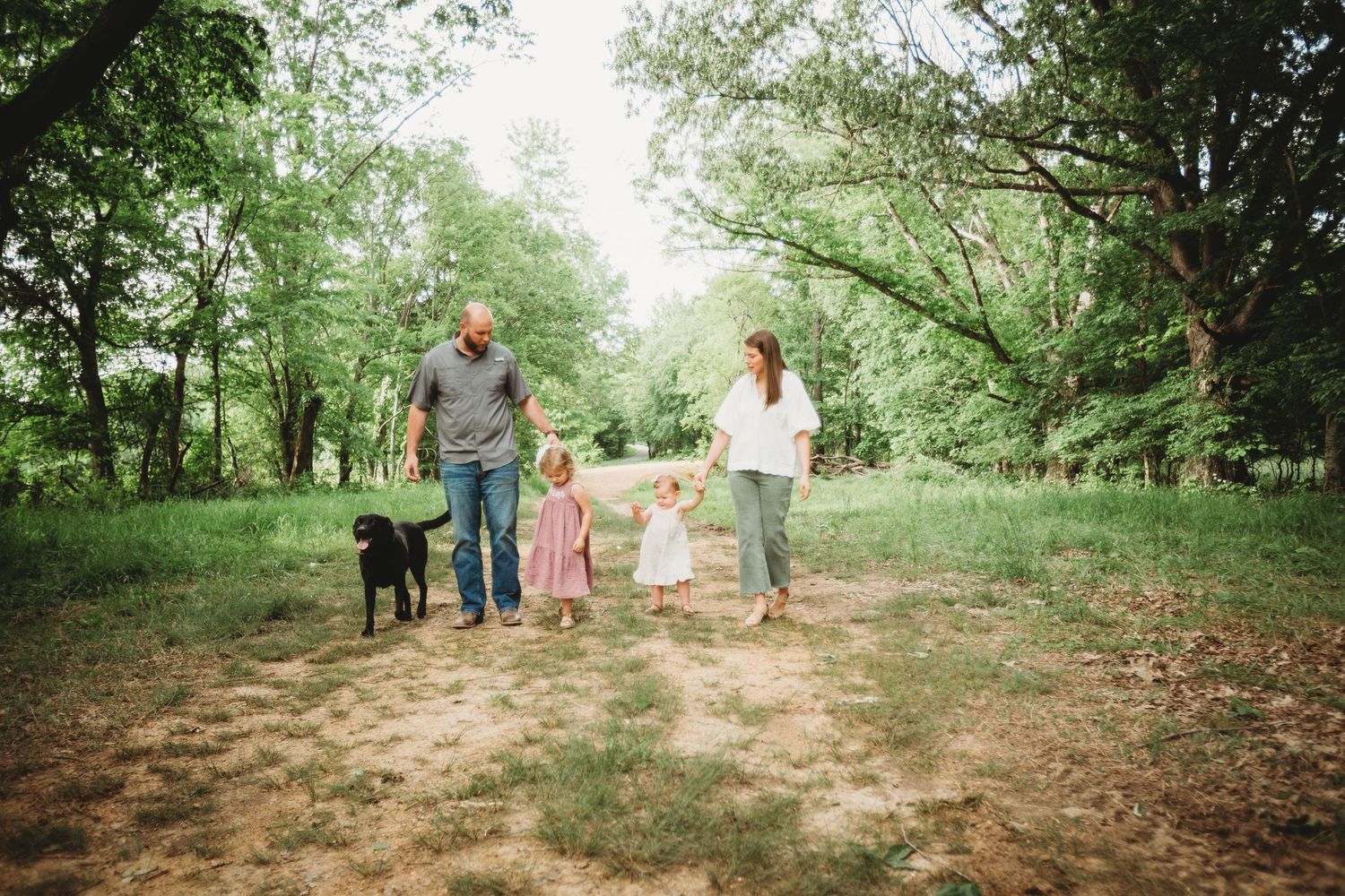 A family walks their dog together on a dirt path through a lush green forest setting.
