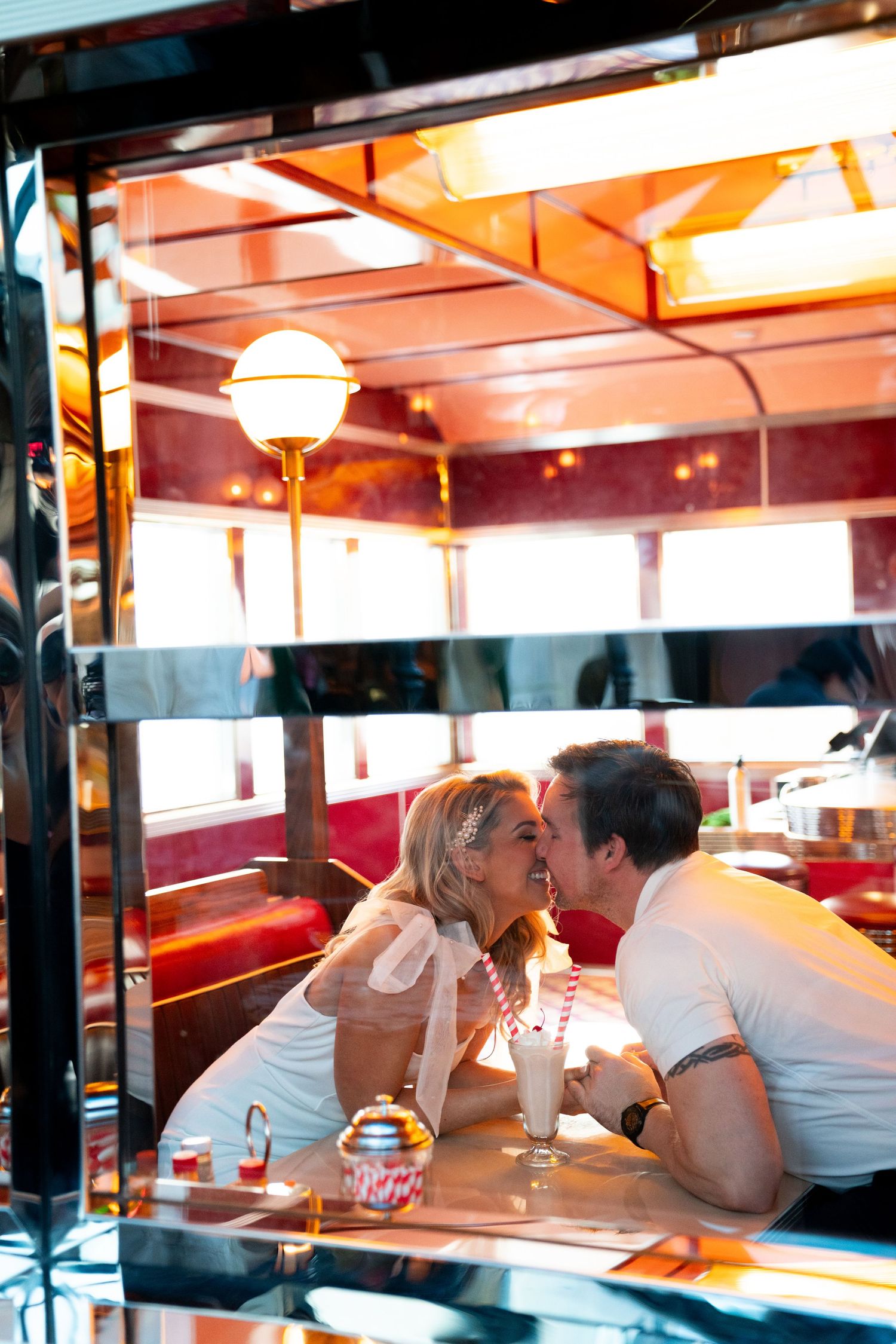 Two people in white outfits share a romantic moment at a retro-style diner booth with red seating and ambient lighting.