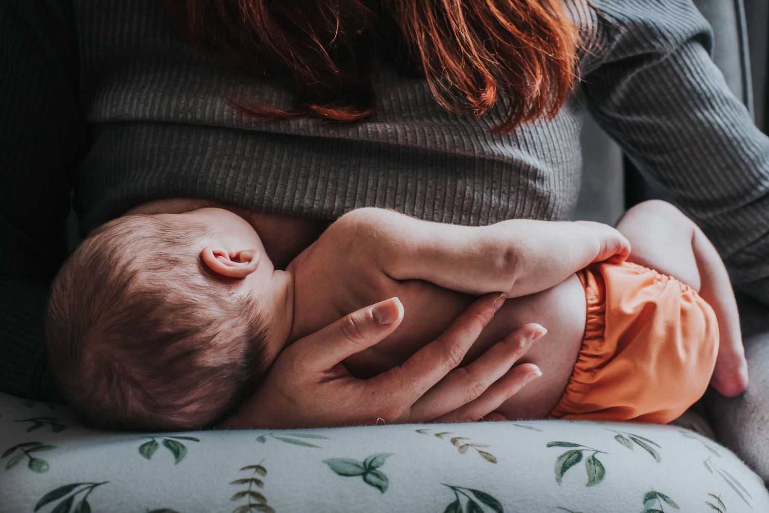 A baby wearing an orange diaper lies cradled in mother's arms, breastfeeding on a floral patterned cushion.
