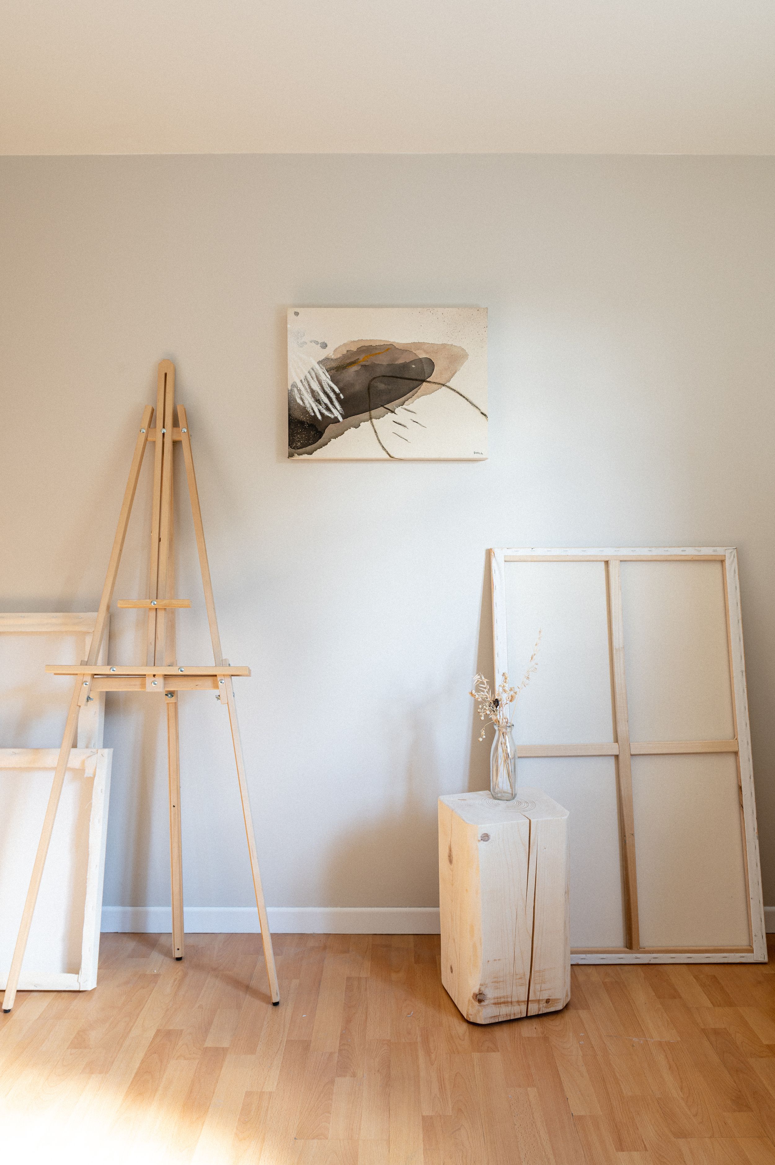 Minimalist art studio corner with wooden easel, framed artwork, and vintage window frame against light gray wall.