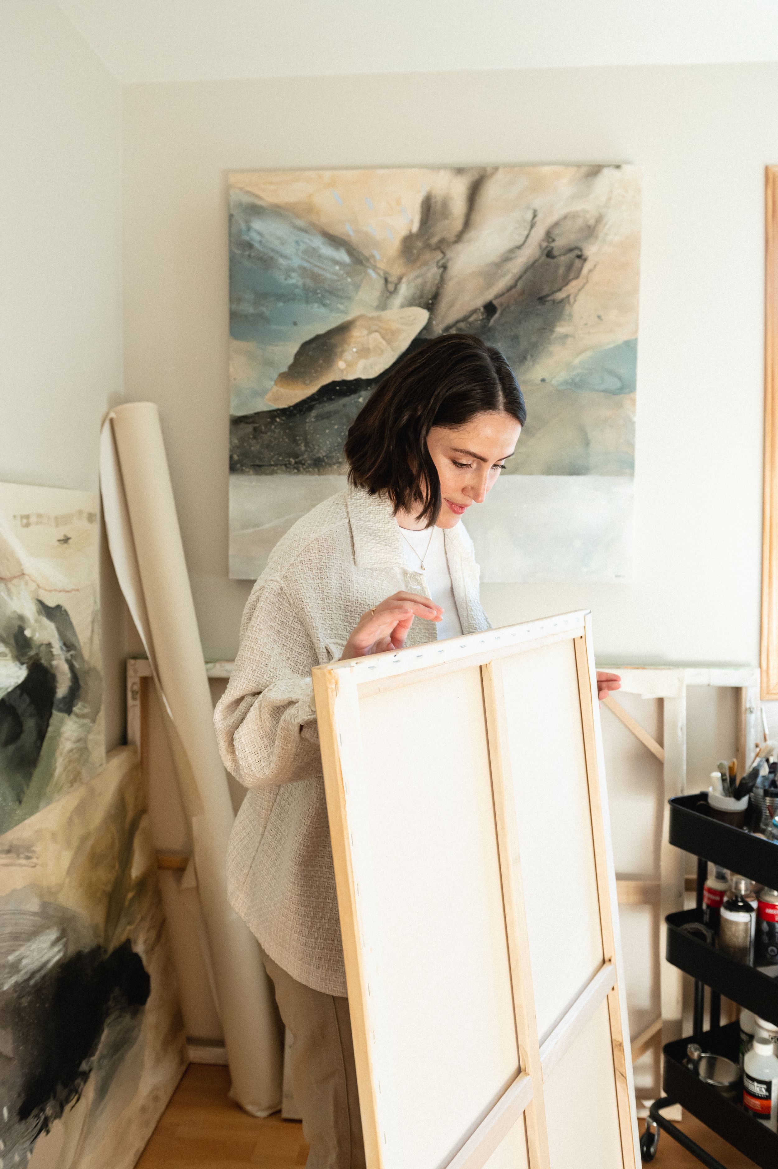 Artist works at easel in bright studio space with abstract landscape artwork displayed on wall.
