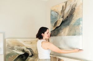Artist stands in front of large abstract artwork while extending arms to gesture at the neutral-toned painting.