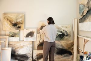 Artist works on abstract paintings displayed against a white wall in a bright studio space.