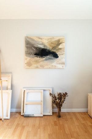 Abstract wall art in muted tones hangs above empty frames and dried flowers on hardwood floor against gray wall.