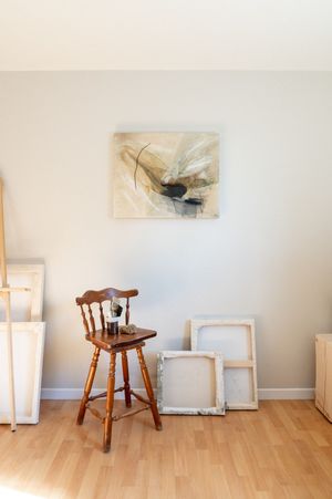 Wooden stool sits against white wall with artwork and light wood flooring in minimalist interior space.