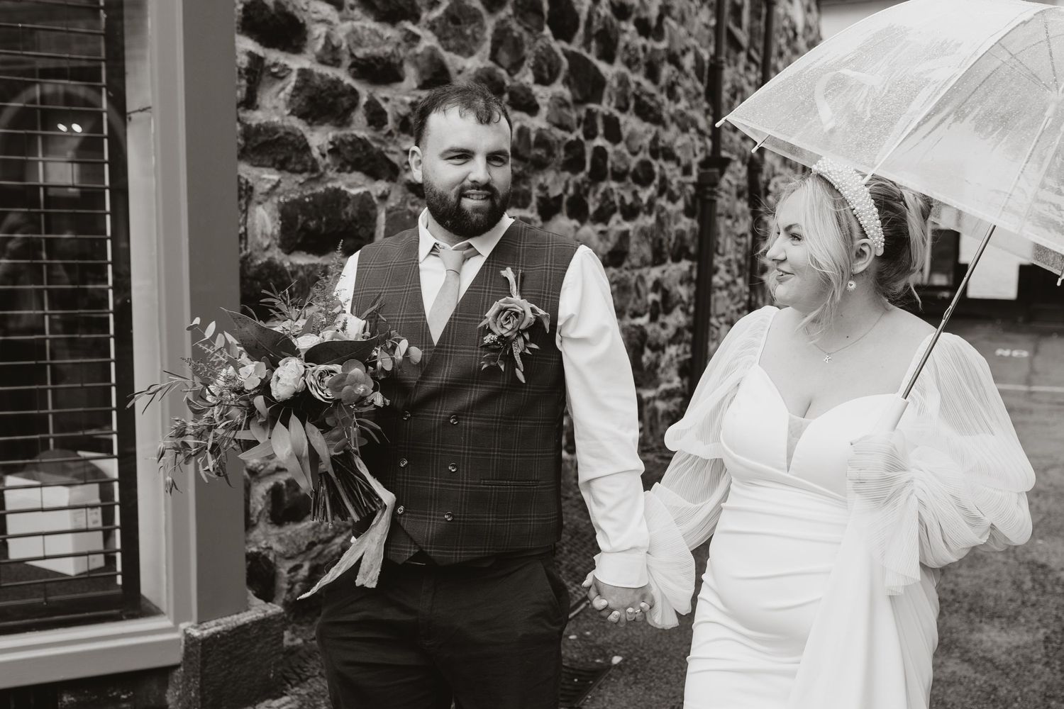 A bride with a parasol and groom in a vest hold hands while walking together near a stone wall.