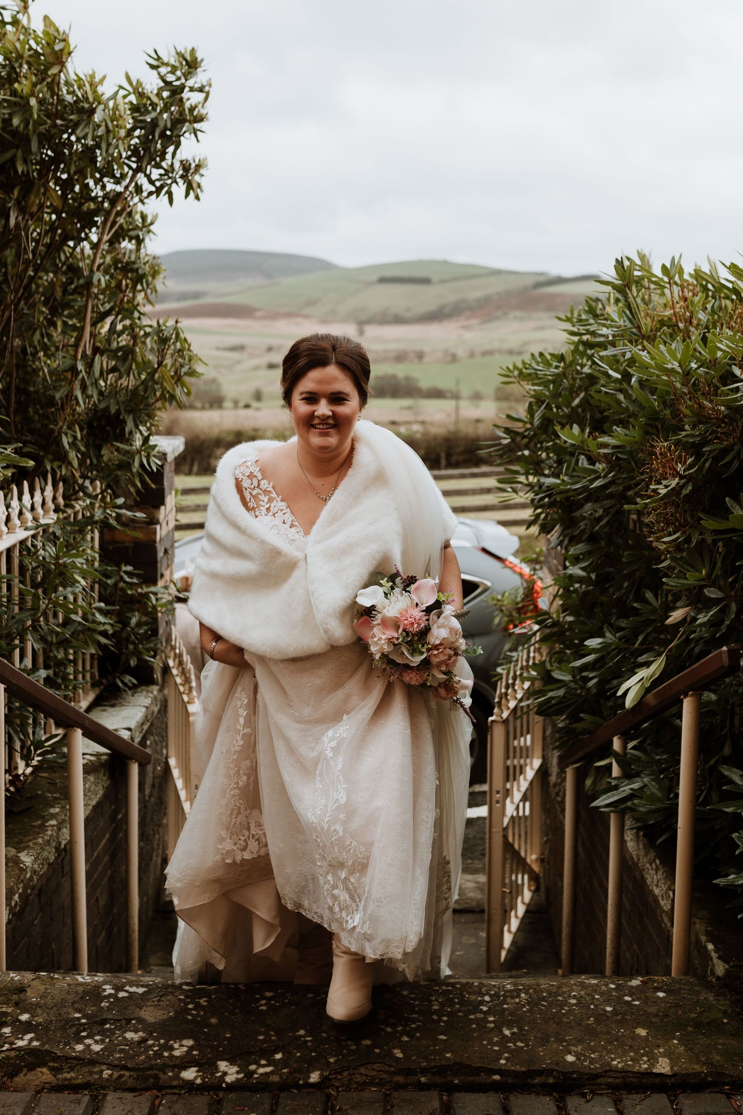 A white lace wedding dress adorned with a cozy fur wrap in a rustic outdoor setting with rolling hills in the background.