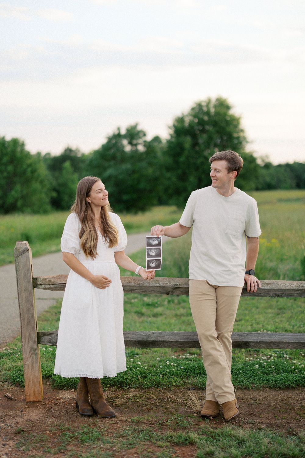 A couple in white and khaki outfits stands by a rustic wooden fence in a pastoral countryside setting at dusk.