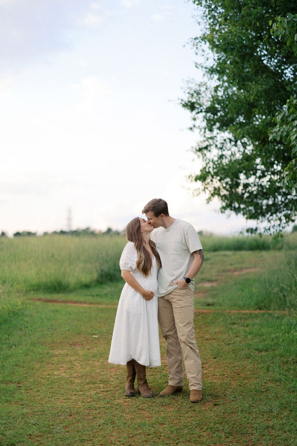 A couple shares a romantic moment on a dirt path in a green field with trees in the background.