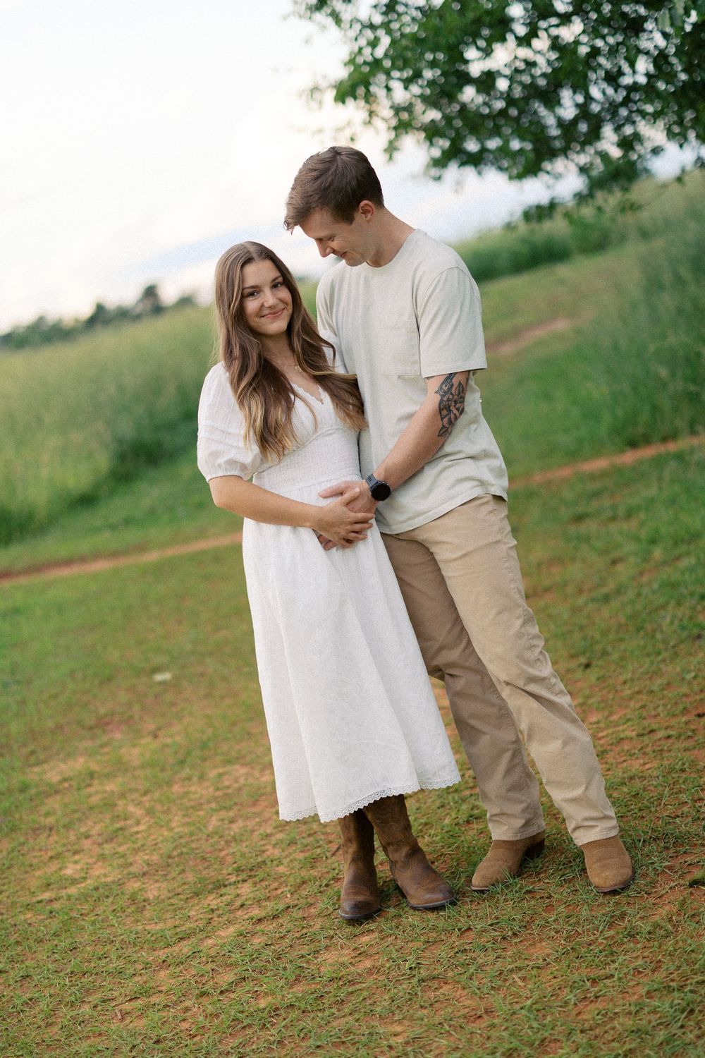 Couple in casual white outfits embrace on grassy hillside during maternity photoshoot outdoors.