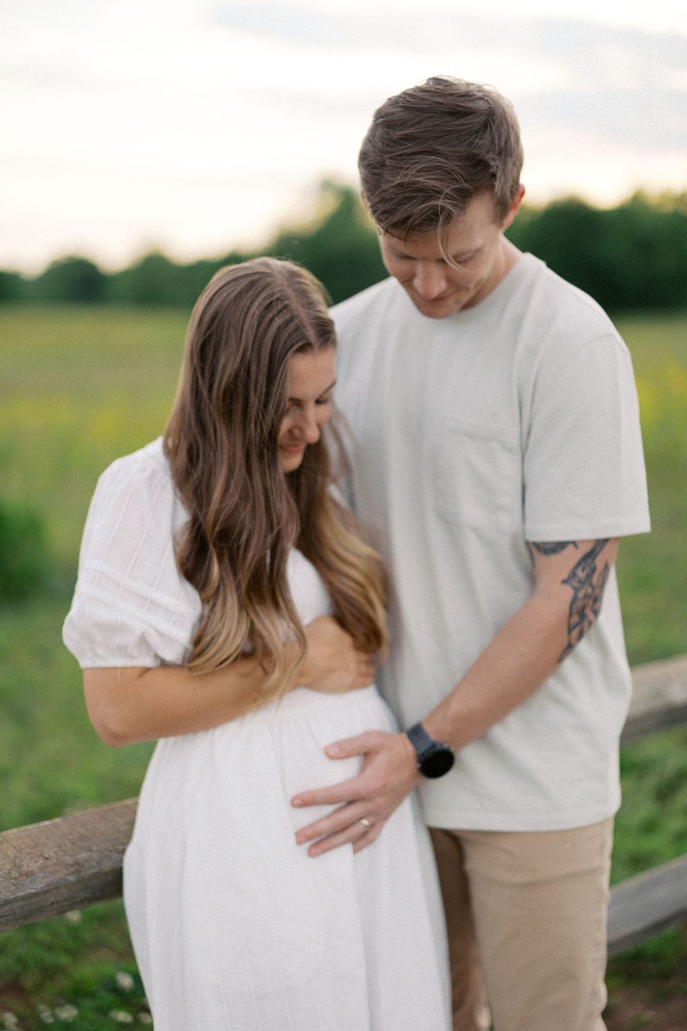 Couple in white outfits embrace and smile while standing in a grassy field during sunset.