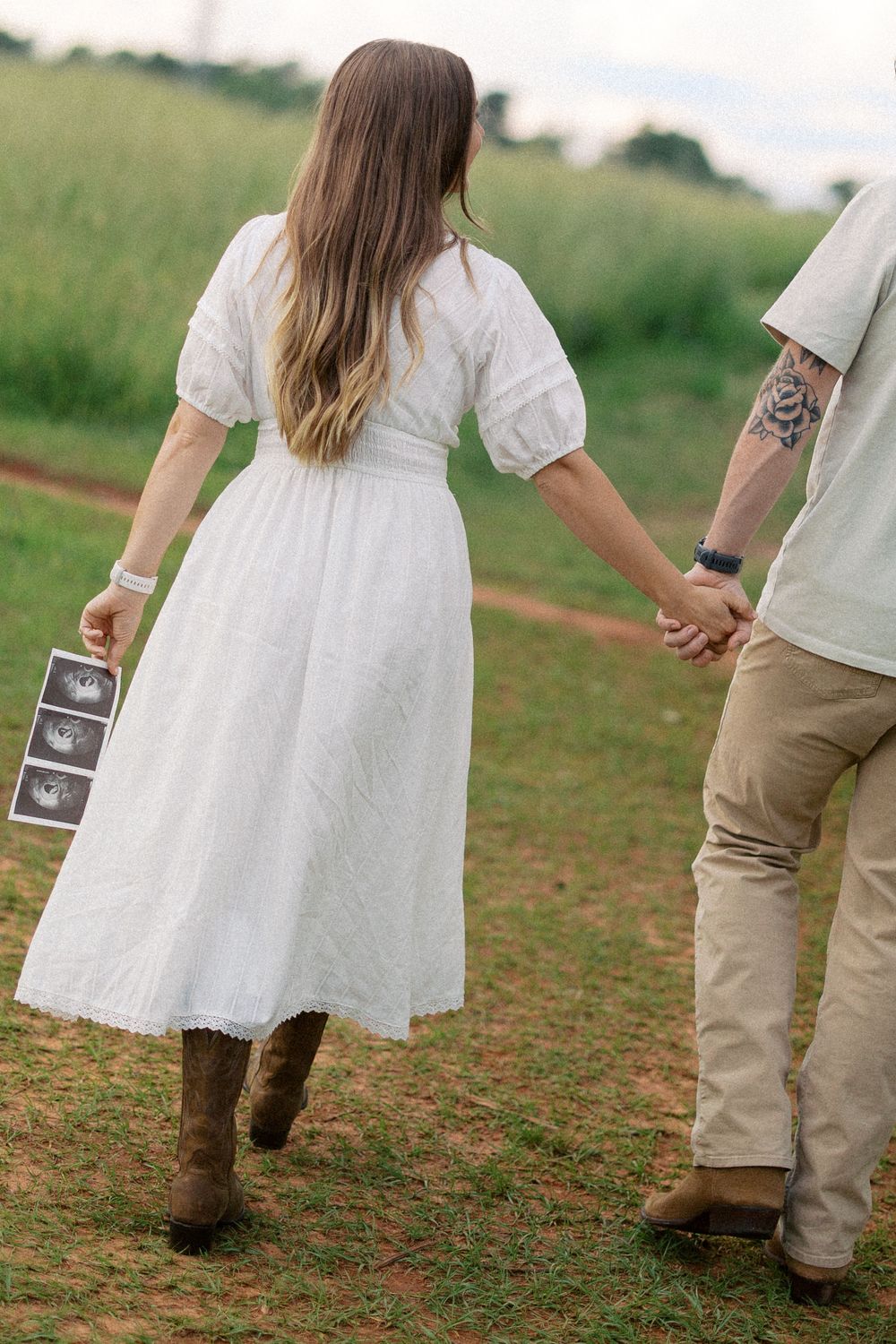A couple holding hands in a field as they walk, with a sonogram photo, wearing white dress and brown boots.