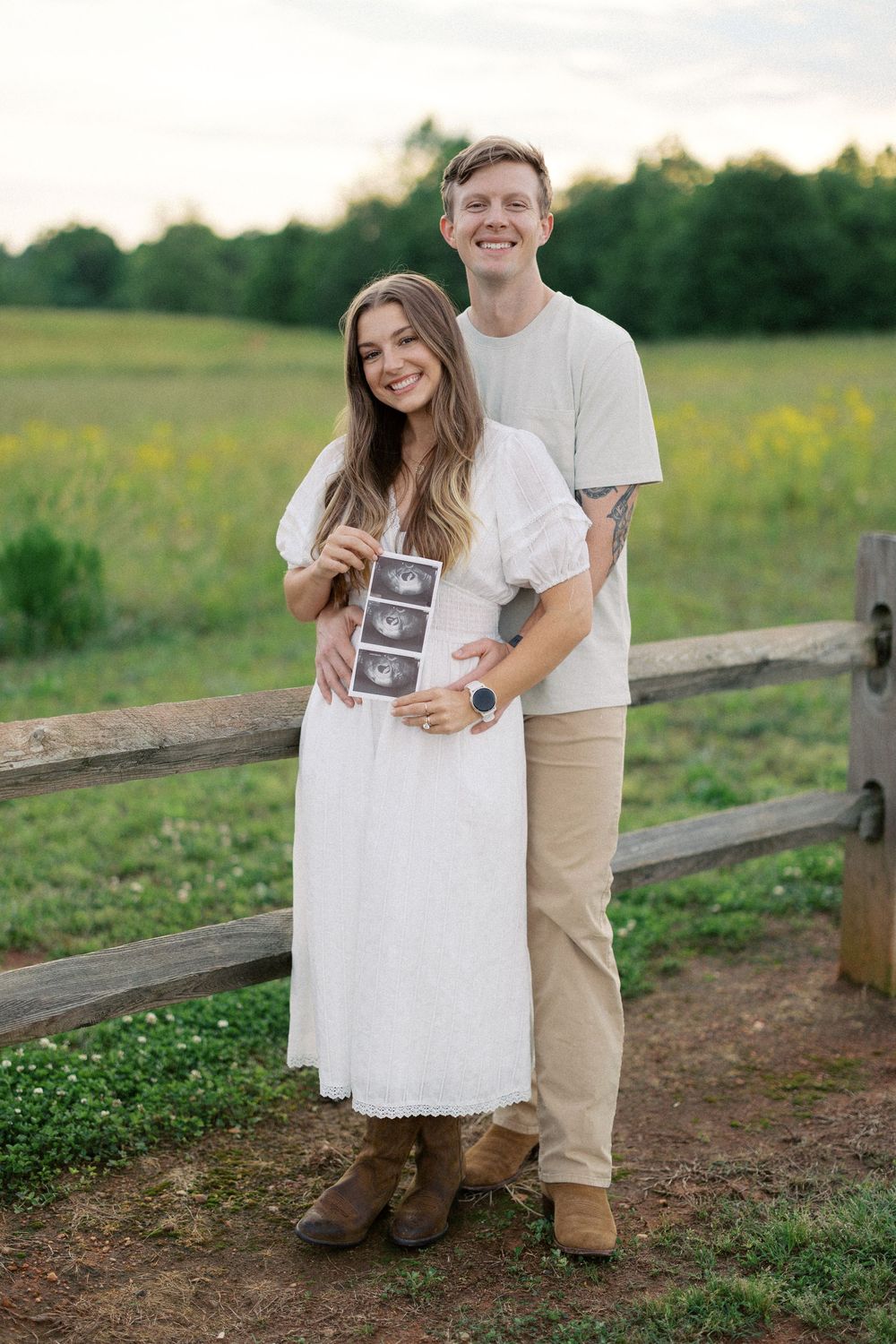 A couple stands together by a wooden fence in a field, holding ultrasound photos while dressed in casual white outfits.