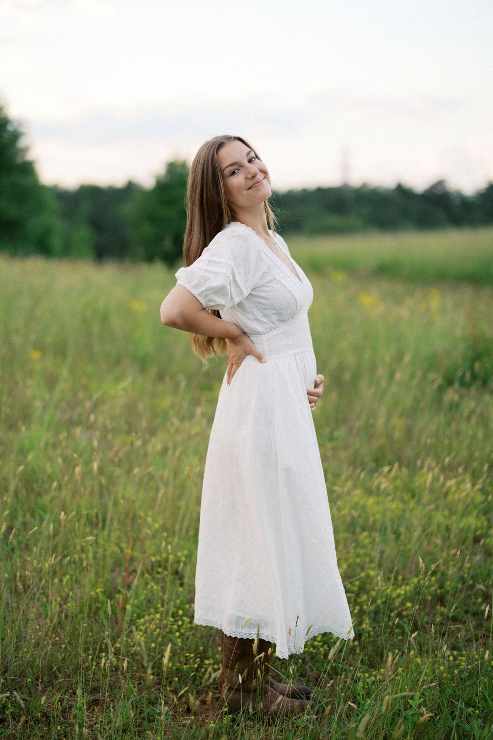 A woman in a flowing white dress stands in a sunlit meadow with tall grass and trees in the background.