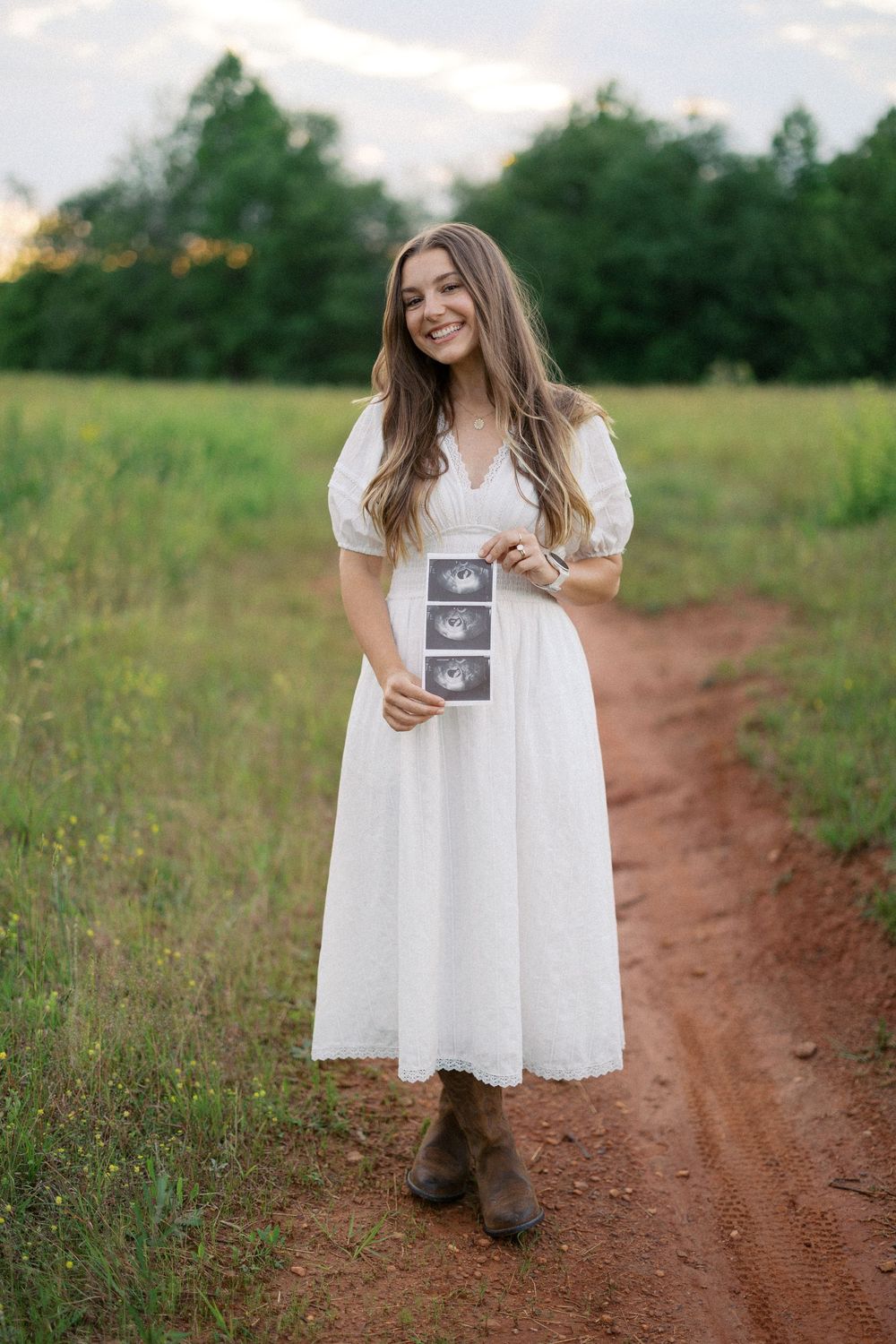 A joyful moment captured on a dirt path as someone in a white dress holds ultrasound photos in a rustic field setting.