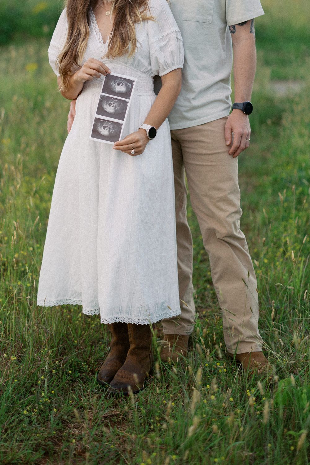 Couple standing in grassy field holding ultrasound photos, she wearing white dress and he in casual attire.