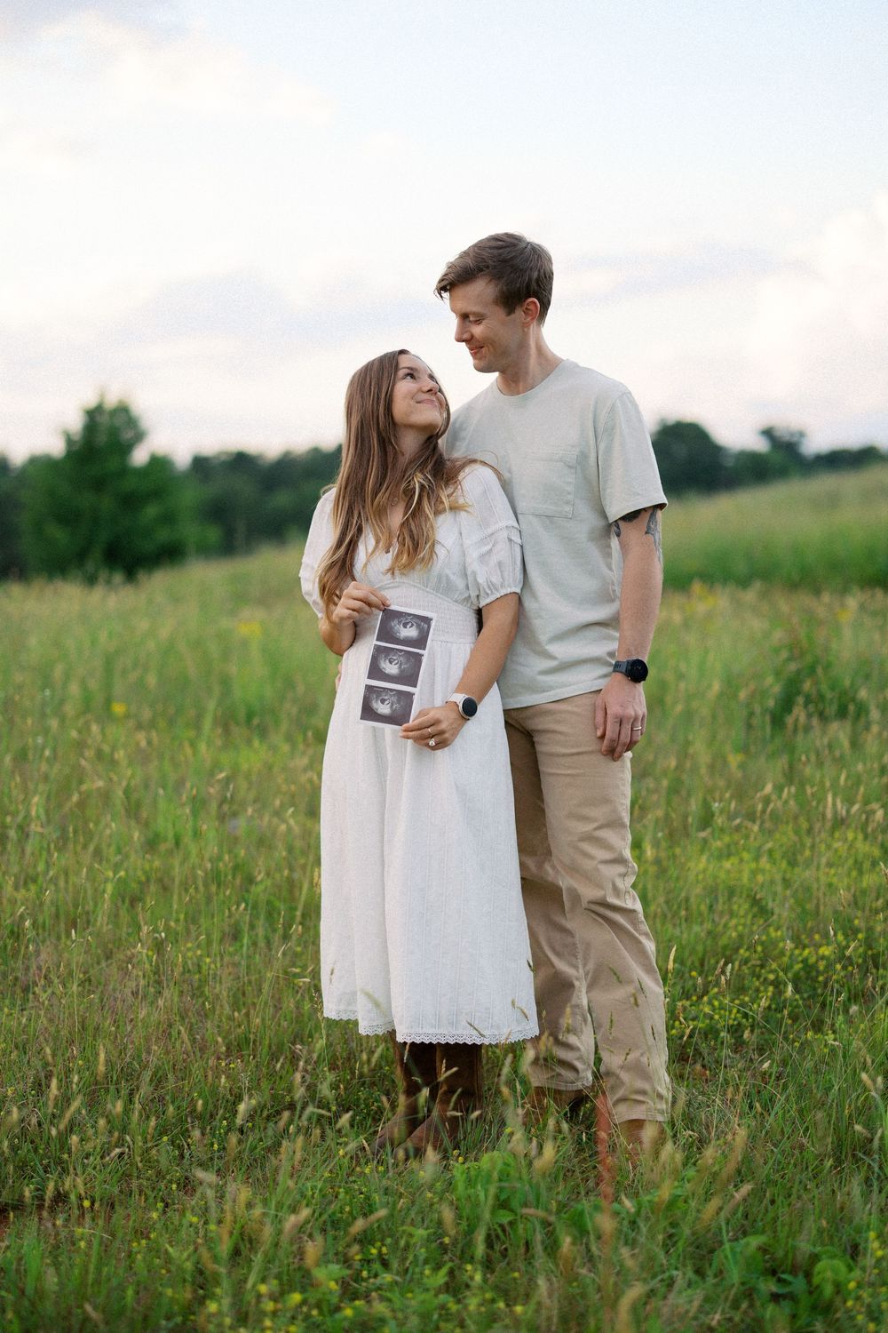Couple embraces in a sunny meadow while holding ultrasound photos.