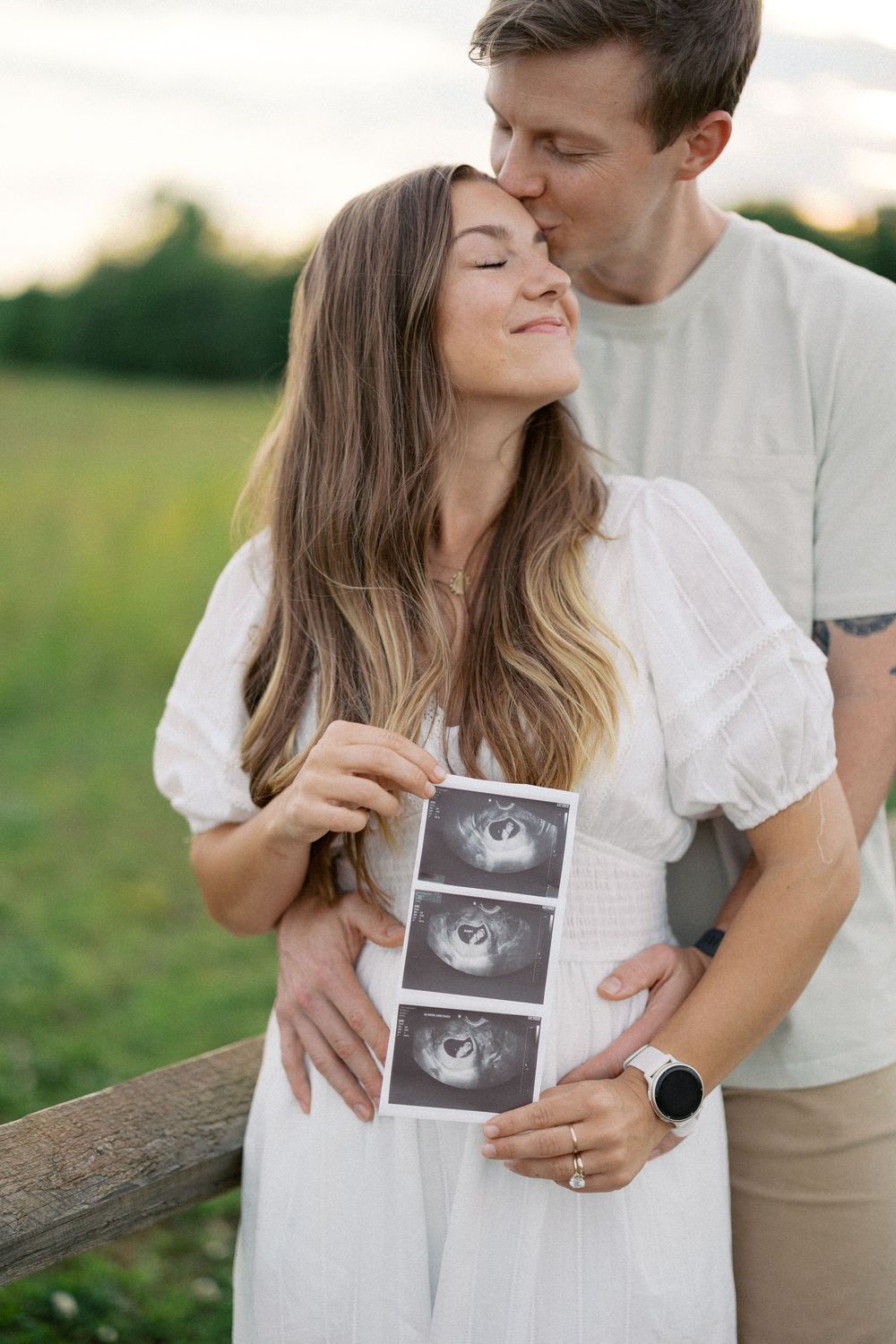 Couple holding ultrasound photos outdoors in a field while sharing an intimate romantic moment at sunset.