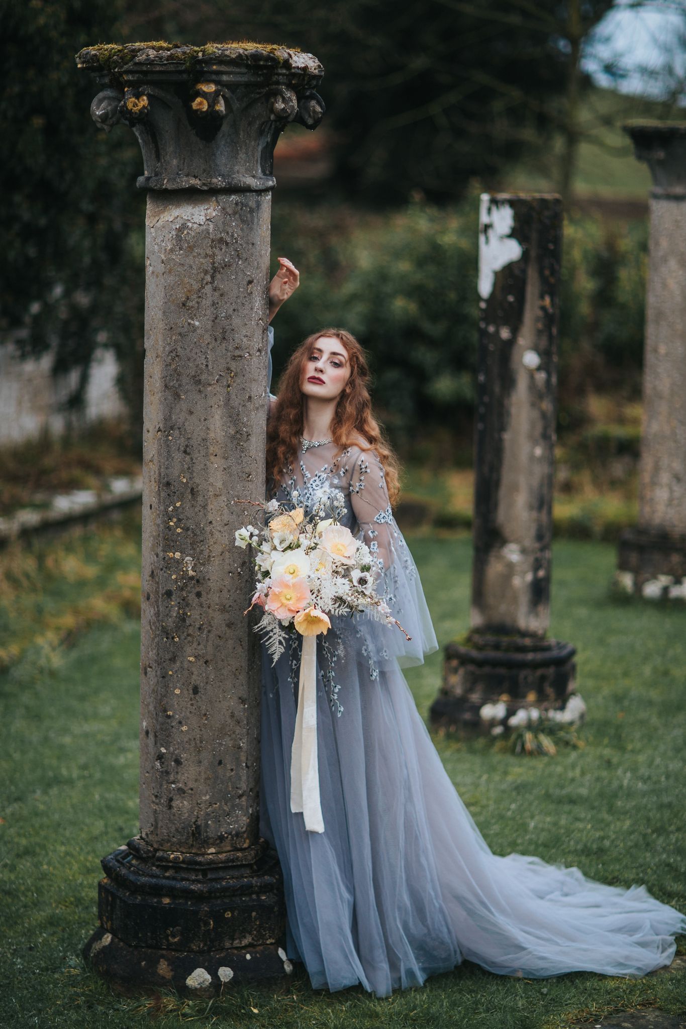 Ethereal wedding scene with flowing grey tulle gown and soft florals against stone pillars in a garden setting.