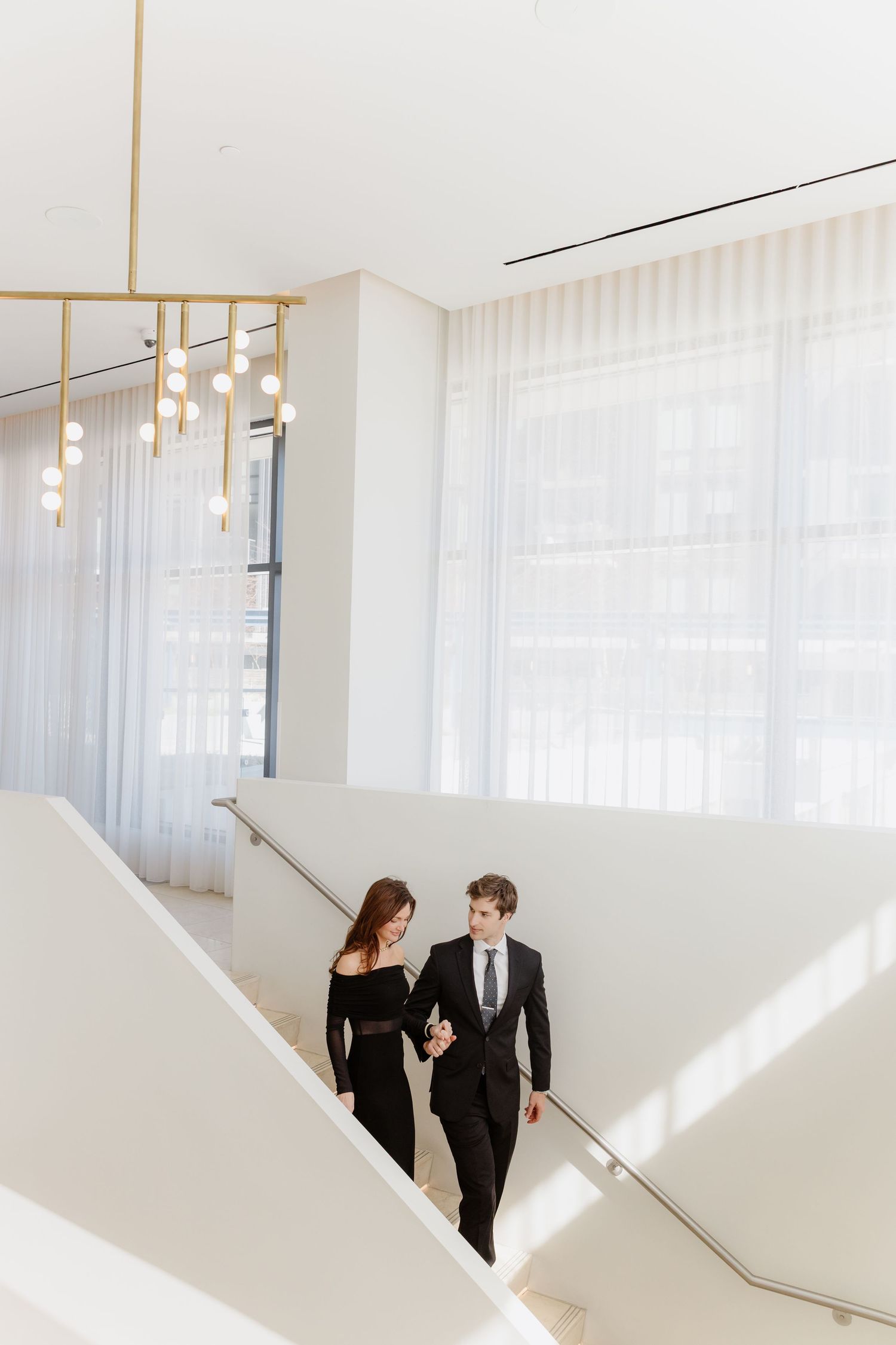 Elegant couple in black formal attire descending white modern staircase with flowing sheer curtains and pendant lights.