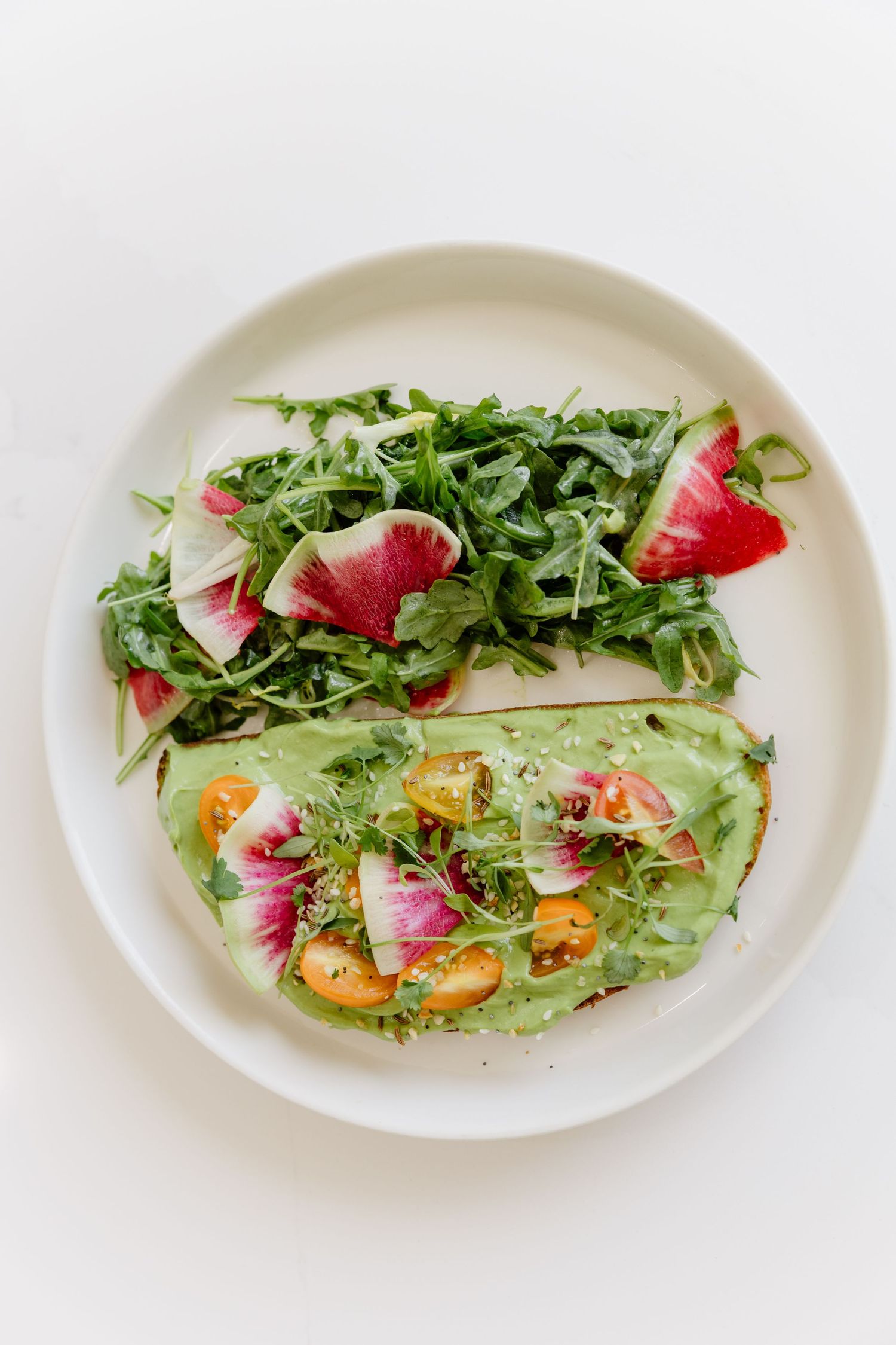 Fresh arugula salad with watermelon radish slices and citrus on white ceramic plate against light background.