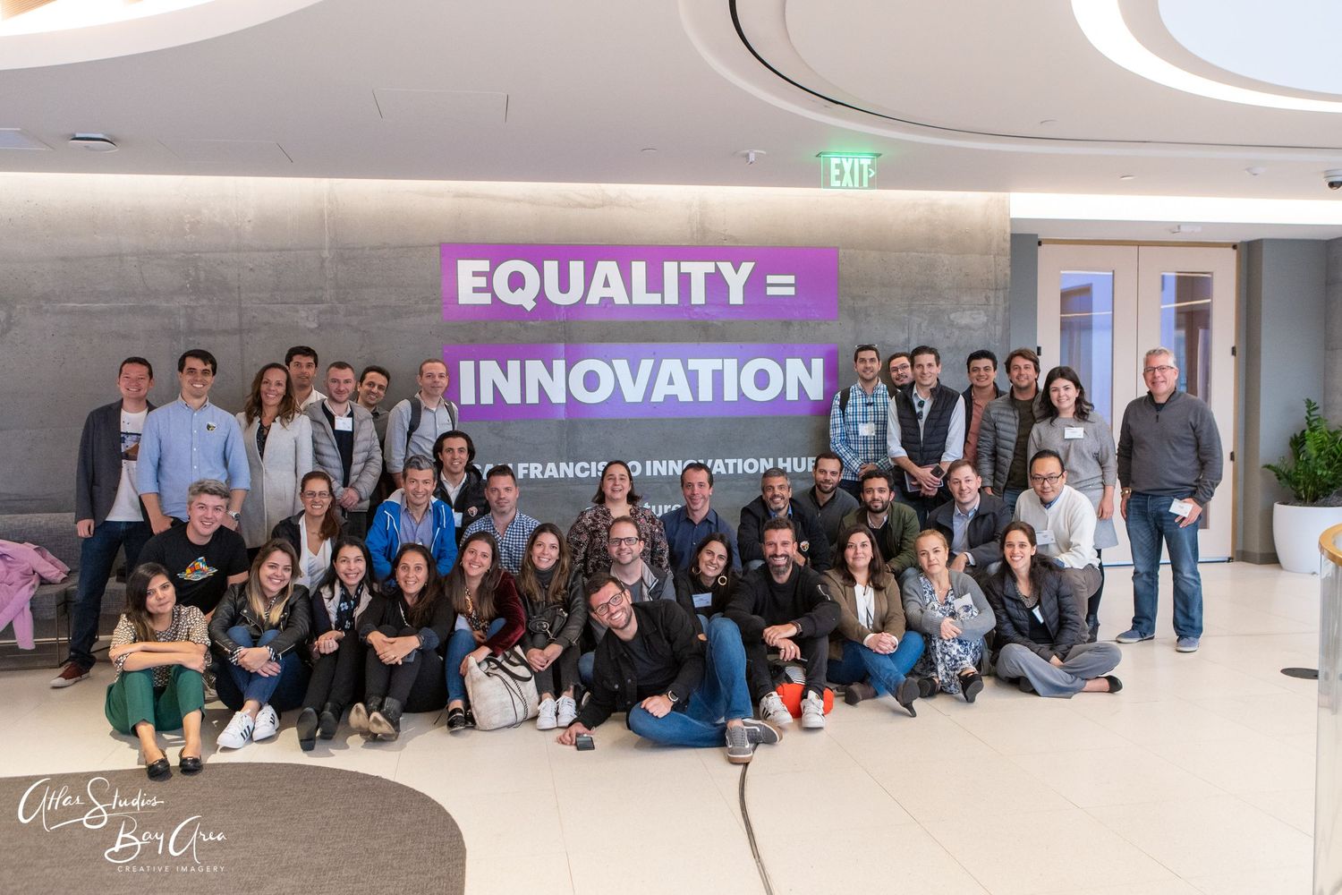 Large group gathered beneath purple 'Equality = Innovation' sign in modern office space.